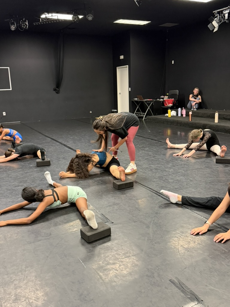 A teacher assisting dancers during a stretch on the studio floor