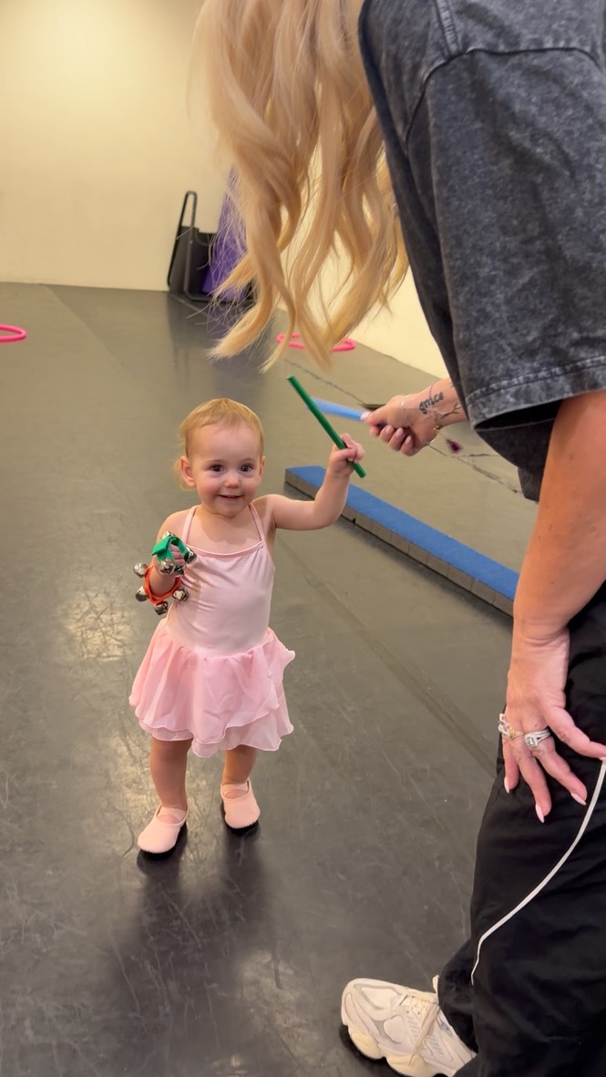 A young dancer holding bells while reaching toward a teacher in the dance studio
