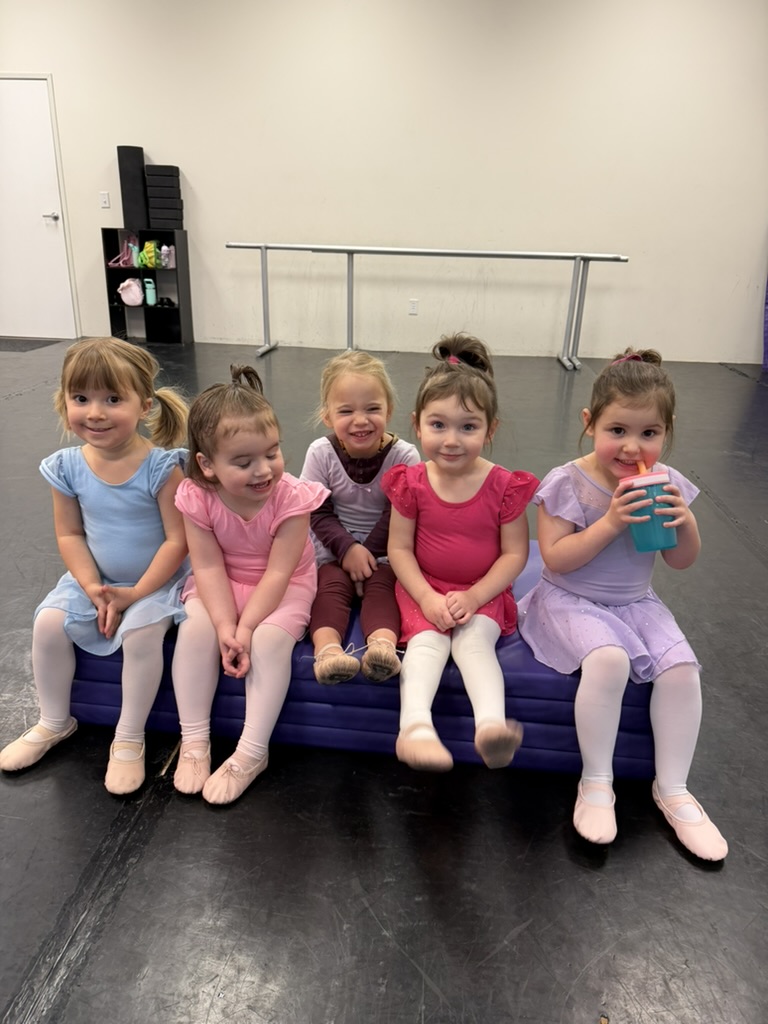 Five young dancers sitting together on a purple mat in the dance studio
